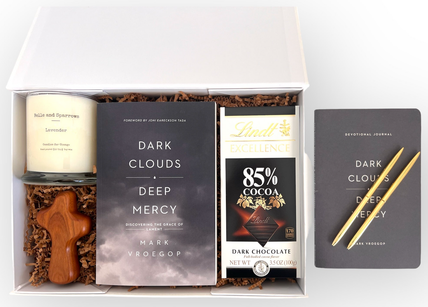 Comfort in Grief gift box with lavender candle by Belle and Sparrows, Deep Clouds Deep Mercy book and devotional journal. Handheld wood cross and two gold ballpoint pens. Lindt 85% Dark Chocolate bar.